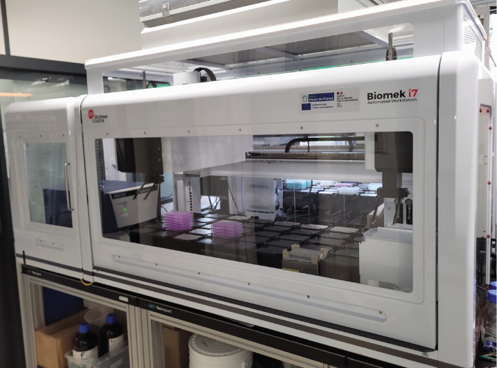 Photographie d'une station de pipetage automatique Biomek i7 à paroie vitrée à travers laquelles le robot de pipetage est visible. Image clicable donnant accès au descriptif des stations de pipetage BiomeK i7.